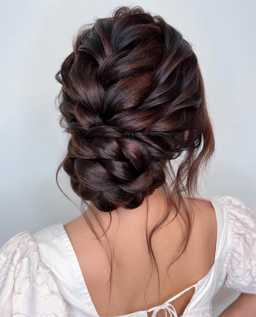 Full brunette braided low bun with wide woven sections and loose neck tendrils