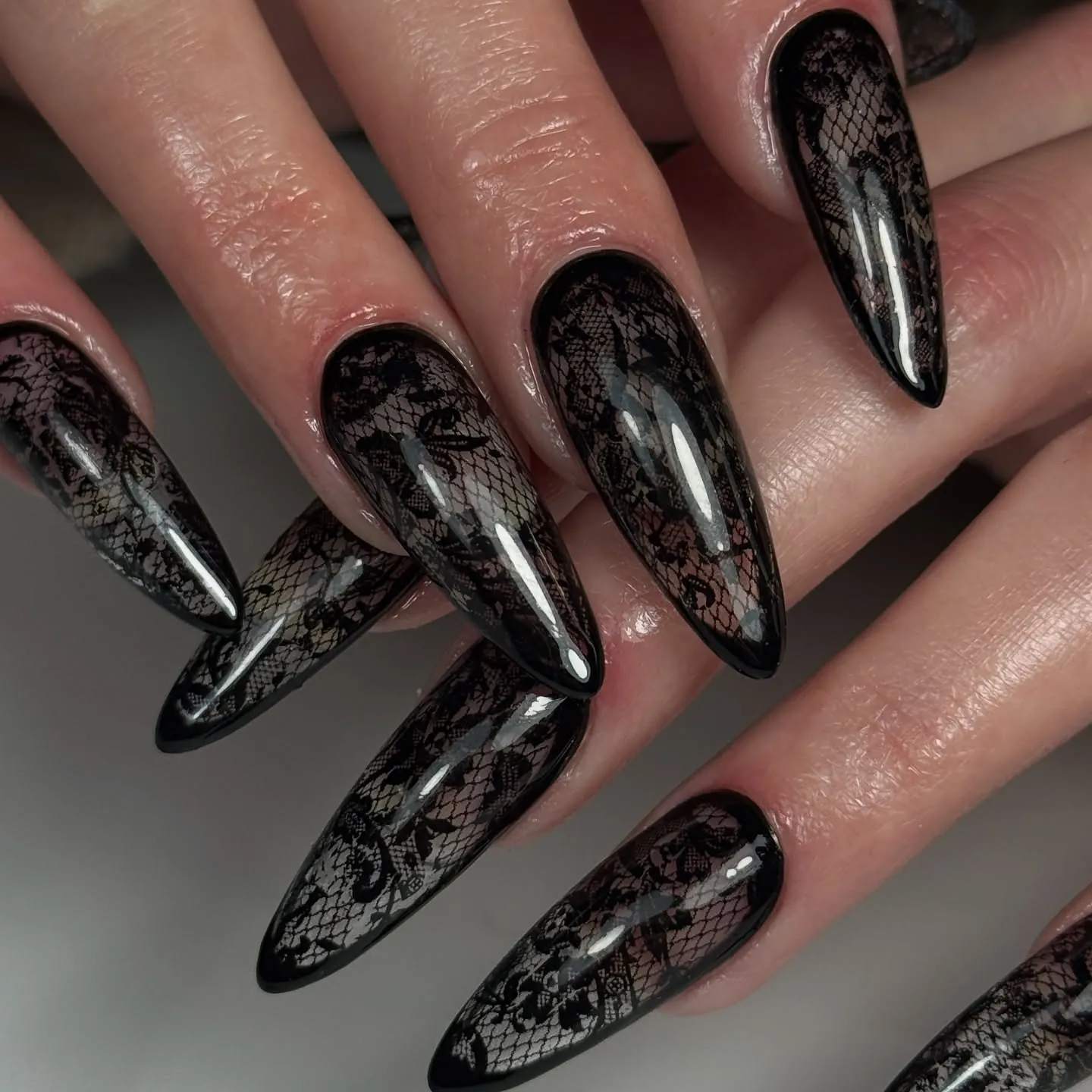 Long almond nails with black floral lace patterns over sheer nude bases