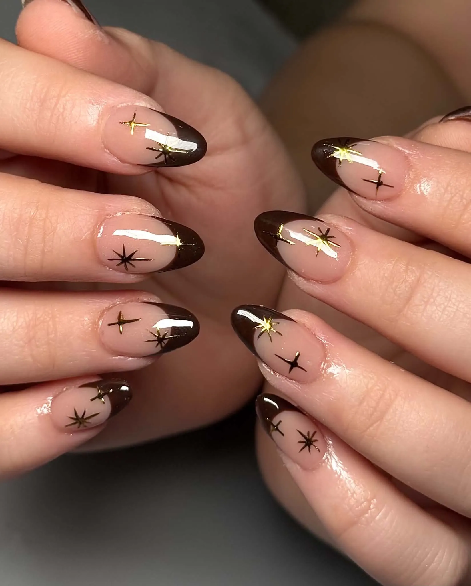 Nude almond nails with dark brown pointed tips and gold starbursts