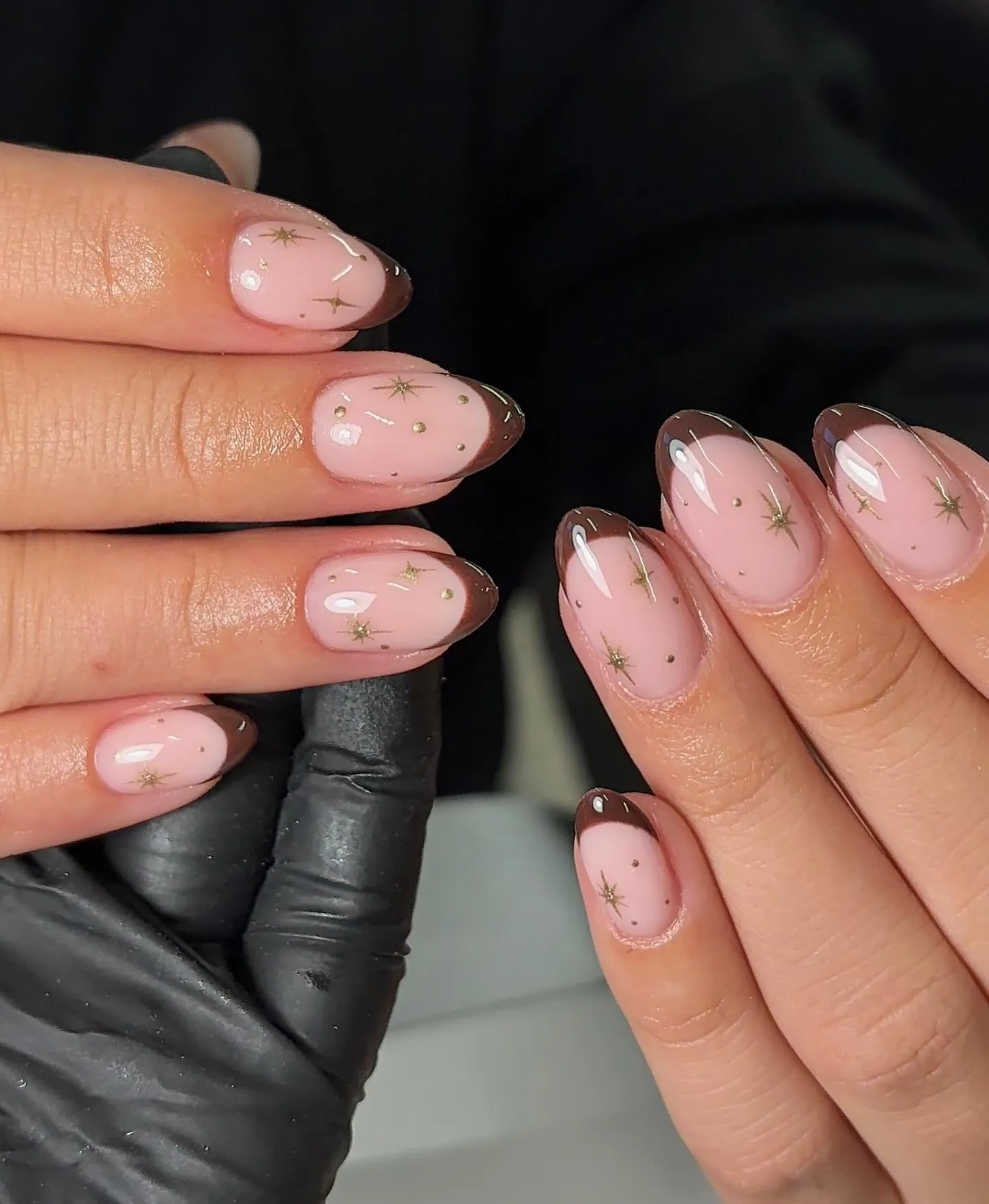 Soft pink almond nails with dark brown curved tips and fine gold starbursts