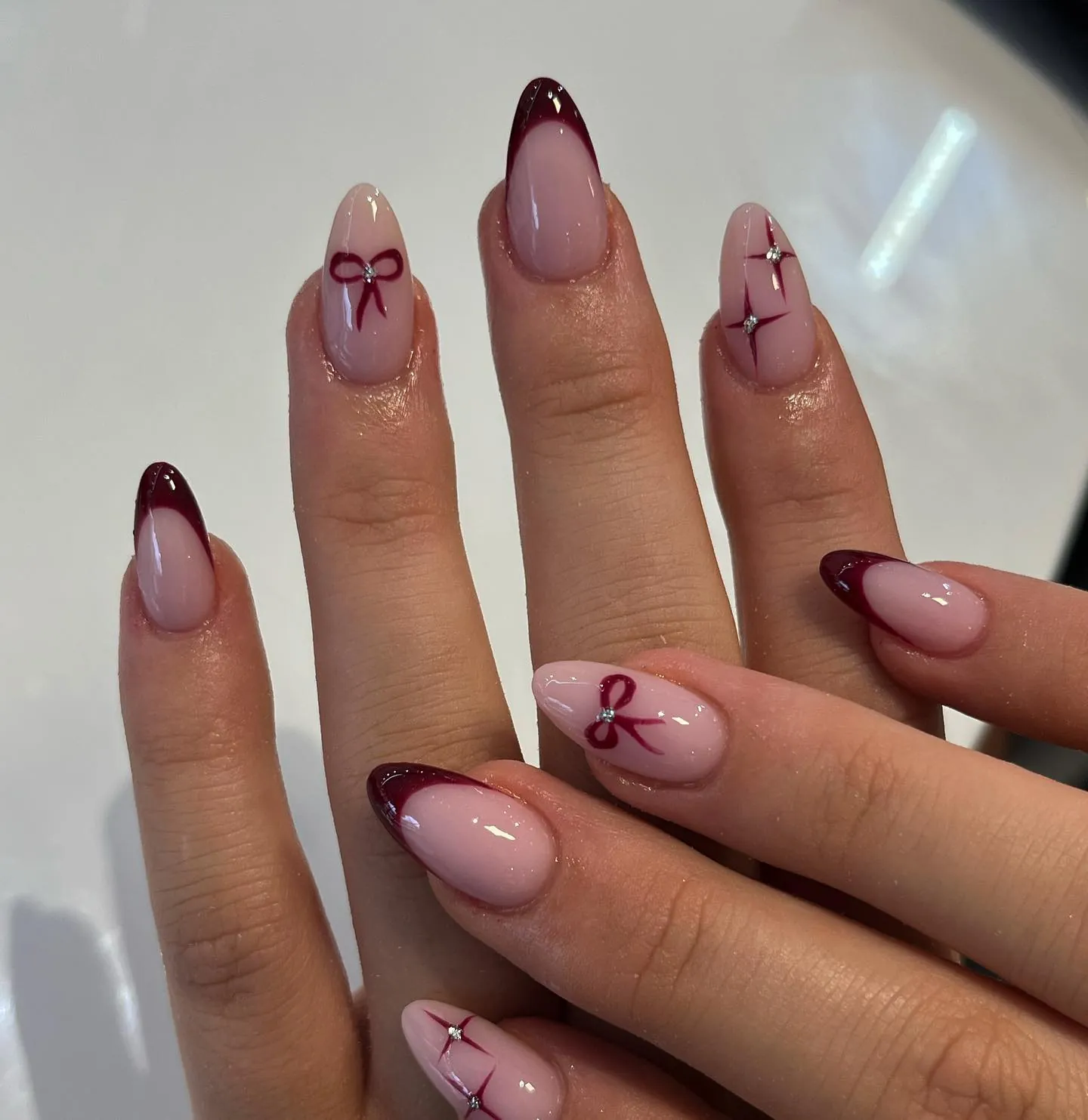 Nude almond nails with burgundy French tips and fine bow artwork