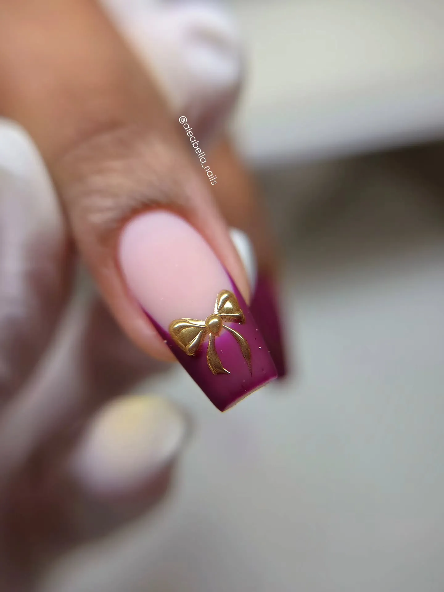 Pink square nail with a burgundy tip and raised gold bow
