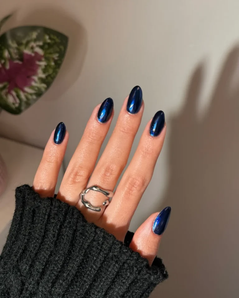 Glossy dark blue almond-shaped nails with elegant navy polish and a minimalist accent nail, styled with silver rings for a chic manicure look.