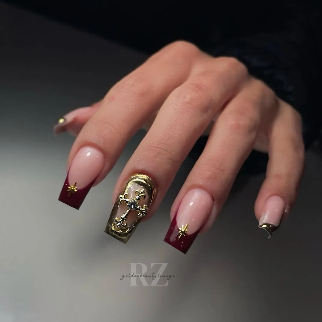 Square burgundy French nails with gold stars and an ornate raised cross accent